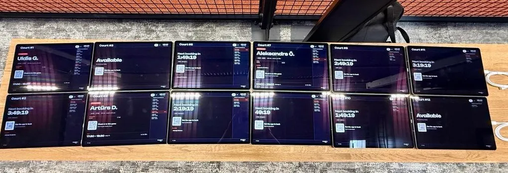 Twelve court tablets laid out on a bench at the venue, each showing its live court screen with booking status and countdown